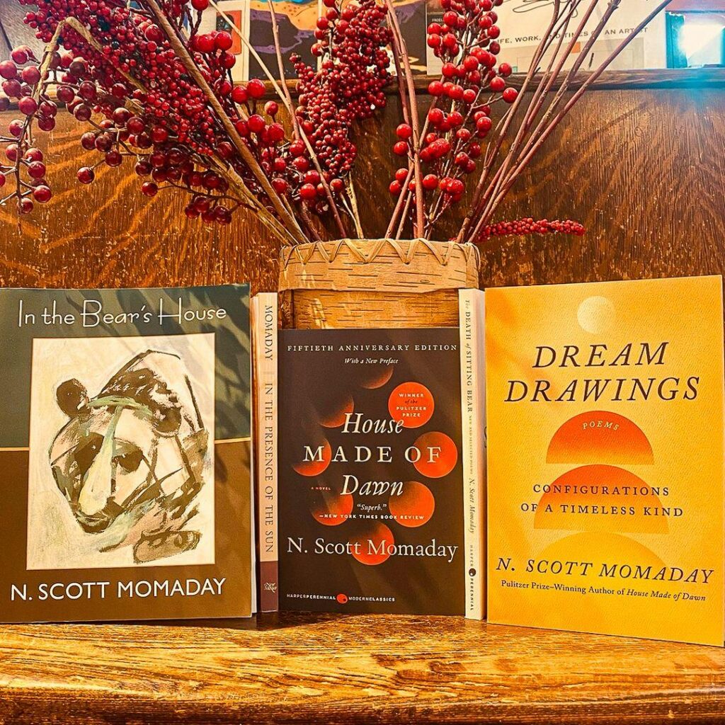 Three books by n. scott momaday displayed on a wooden surface, flanked by red berries and a woven basket.