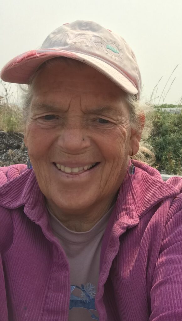 Elderly woman smiling at the camera, wearing a pink baseball cap and pink jacket, outdoors with a foggy background.