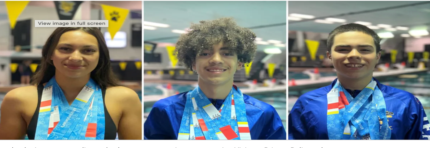 Three young athletes wearing swim team uniforms with medals, smiling in an indoor pool area.