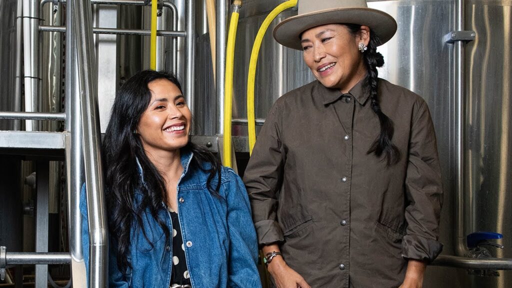 Two women smiling and standing in a brewery with stainless steel equipment in the background. one wears a denim jacket and the other a black shirt and hat.