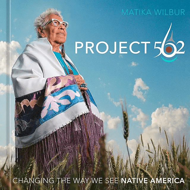 Elderly native american woman in traditional clothing stands in a field under a blue sky, with text "matika wilbur project 562 changing the way we see native america.