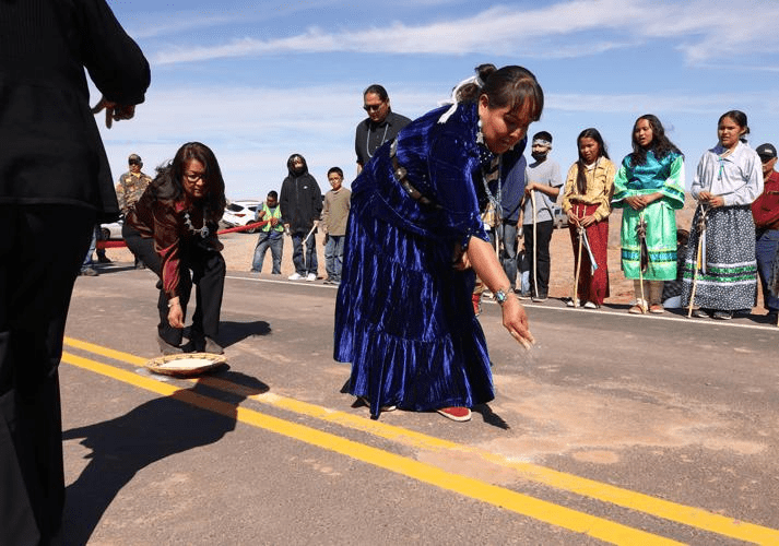 Two women in traditional dress participate in a cultural ceremony on a road, surrounded by a group of onlookers.