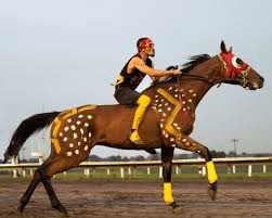 A jockey in red and yellow gear riding a brown horse with decorative spots and matching tack on a racetrack.