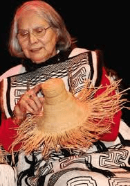 Elderly woman in traditional attire weaving a straw hat, sitting on a stage focused on her craft.