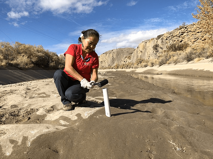 A woman in a red shirt and gloves collects soil samples by a river using a small shovel and a pvc pipe.